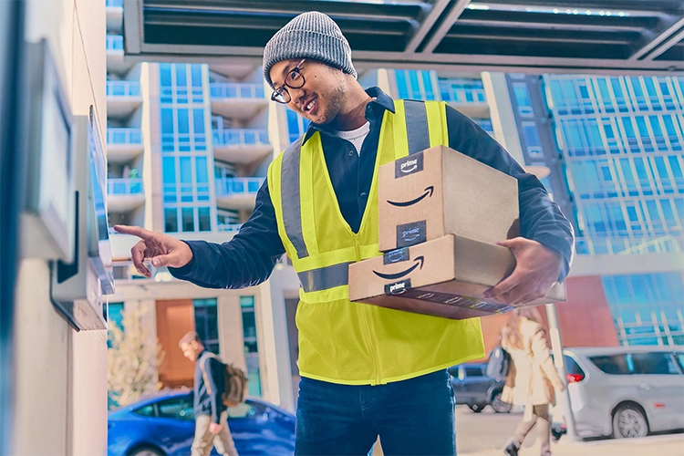 Man in yellow vest, winter hat and glasses holding Amazon packages and entering an apartment building on an urban street