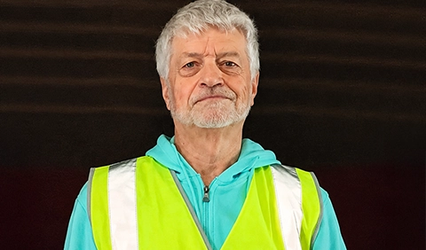 Man with gray hair and a gray beard wearing a yellow vest looking at the camera